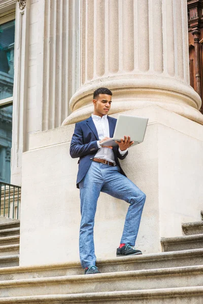 Afrikalı Amerikalı üniversite öğrencisi New York 'ta okuyor. Mavi ceketli, beyaz gömlekli, gri pantolonlu, spor ayakkabılı, genç, ofis binasının dışındaki sütuna karşı merdivenlerde duruyor, dizüstü bilgisayarda çalışıyor..