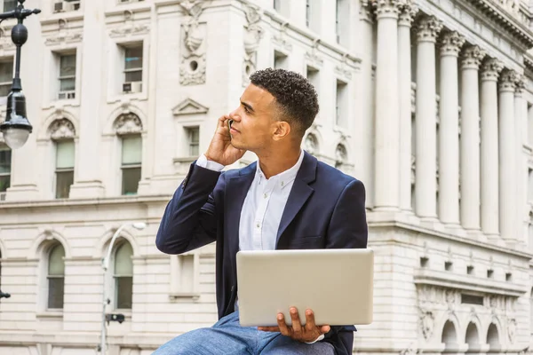 City life. African American college student studies in New York. Young man sits by vintage office building on street, reads, works on laptop computer, looks up, talks on cell phone.