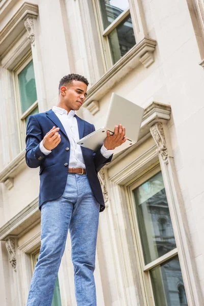 Way to Success. African American male college student studies in New York. Wearing blue blazer, white shirt, young man stands outside vintage office building, looks down, works on laptop computer.