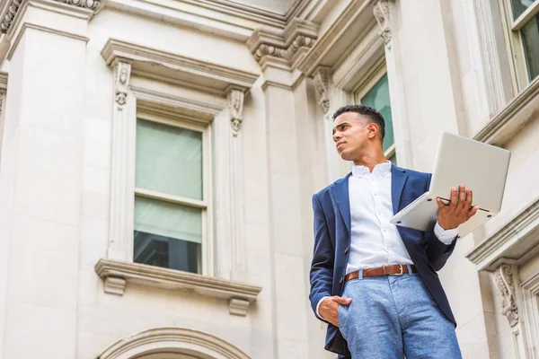 Way to Success. African American male college student studies in New York. Wearing blue blazer, white shirt,  man stands outside vintage office building, works on laptop computer, looks up, thinks.