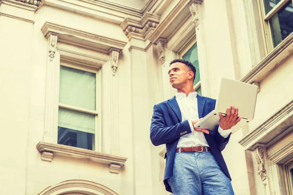 Way to Success. African American male college student studies in New York. Wearing blue blazer, white shirt,  man stands outside vintage office building, works on laptop computer, looks up, thinks.