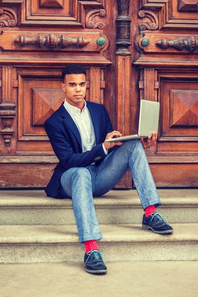 Başarının yolu. Mavi ceketli, gri pantolonlu, spor ayakkabılı, Afrikalı Amerikalı üniversite öğrencisi New York 'ta kampüsün klasik ofis kapısında oturuyor, dizüstü bilgisayarla çalışıyor.. 