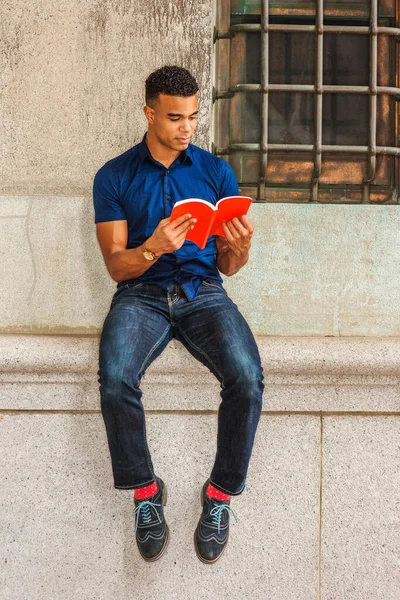 African American College Student studies in New York. Wearing blue short sleeve shirt, jeans, red socks, sneakers, young man sits against vintage wall on street, reads red book.