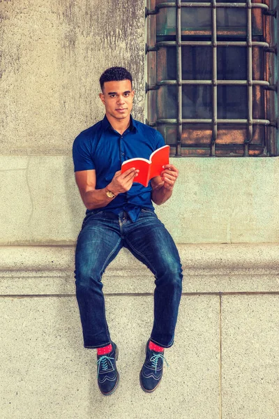 African American College Student studies in New York. Wearing blue short sleeve shirt, jeans, red socks, sneakers, young man sits against vintage wall on street, reads red book.