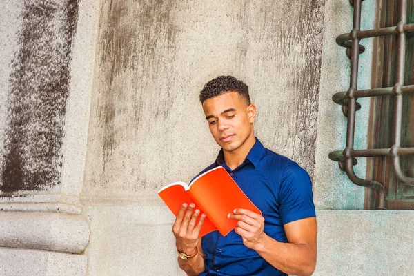 I love reading book. African American College Student studies in New York. Young man wearing blue short sleeve short, sits against vintage wall on campus, looks down, concentratedly reads red book.