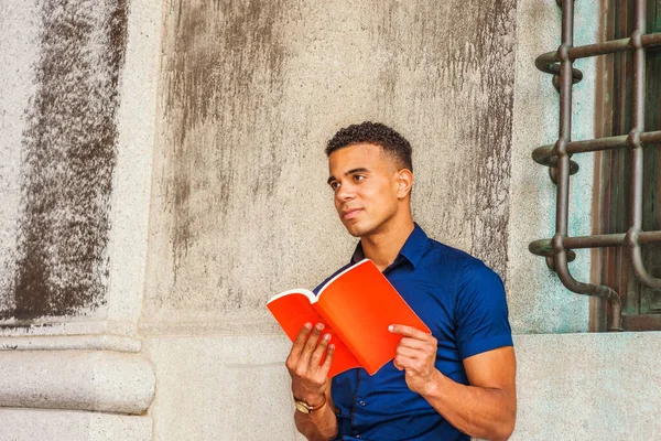 I love reading book. African American College Student studies in New York. Young man wearing blue short sleeve short, sits against vintage wall on campus, reads red book, looks up, seriously thinks.