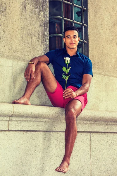 I miss you, wait for you. Wearing blue short sleeve shirt, red shorts, bare foot, African American man sits against vintage wall on street in New York, holds white rose, looks around.