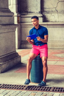 Modern okuma. Mavi kısa kollu gömleği, kırmızı şortu, spor ayakkabısı olan Afro-Amerikan adam New York 'un klasik caddesinde metal sütuna oturmuş, küçük tablet bilgisayar okuyor.. 