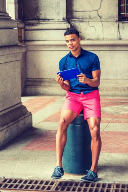 Modern okuma. Mavi kısa kollu gömleği, kırmızı şortu, spor ayakkabısı olan Afro-Amerikan adam New York 'un klasik caddesinde metal sütuna oturmuş, küçük tablet bilgisayar okuyor.. 