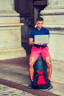 Afro-Amerikan Koleji öğrencisi seyahat ediyor, New York 'ta okuyor. Mavi kısa kollu gömlekli, kırmızı şortlu, sırt çantalı, sokakta oturan, dizüstü bilgisayarda çalışan bir adam.. 