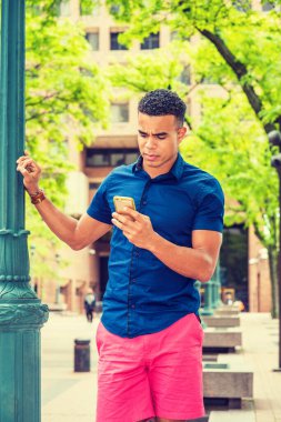 Afro-Amerikan üniversite öğrencisi dışarıda mesaj atıyor. Mavi kısa kollu, kırmızı şortlu, yazın New York 'ta kampüste sokak lambası direğinde duran, cep telefonundan mesajlar okuyan bir adam..