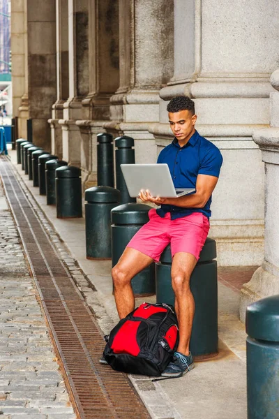 African American College Student travels, studies in New York. Man wearing blue short sleeve shirt, red shorts, with backpack on ground, sits on street, works on laptop computer.