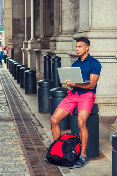 African American College Student travels, studies in New York. Man wearing blue short sleeve shirt, red shorts, with backpack on ground, sits on street, works on laptop computer.