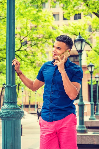 African American college student calling outside, wearing blue short sleeve shirt, red pants, standing by light pole on street on campus in New York in summer, smiling, talking on cell phone.