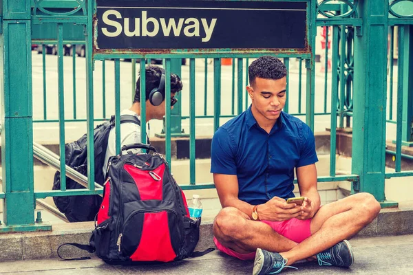 African American college student traveling, studying in New York, wearing blue short sleeve shirt, red shorts, sneakers, bag with bottle water on ground, sitting on street by Subway sign, texting.