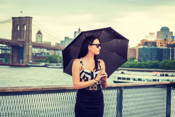 Raining day - grainy, foggy, drizzly feel. American Woman wearing patterned crop bra top, black skirt, sunglasses, holding umbrella, traveling in New York by river. Brooklyn bridge, boat on backgroun