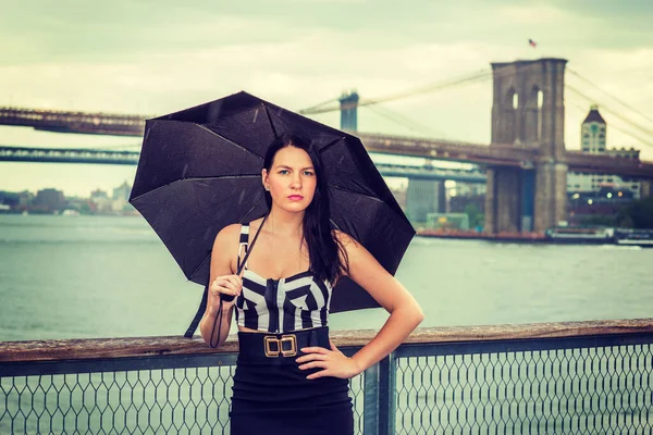 Raining day - grainy, foggy, drizzly feel. Woman wearing black and white patterned crop bra top, black fitted skirt, holding umbrella, traveling in New York. Brooklyn, Manhattan bridges on background