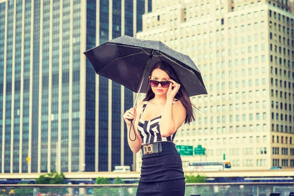 Raining day - grainy, foggy, drizzly feel. American Woman wearing patterned crop bra top, black skirt, sunglasses, holding umbrella, standing in business district in New York, looking over sunglasses
