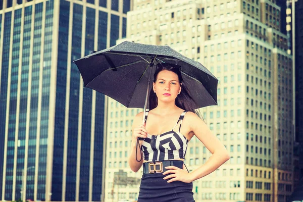 Raining day - grainy, foggy, drizzly feel. American Woman wearing patterned crop bra top, black skirt, sunglasses on head, holding umbrella, standing in business district in New York, looking at you