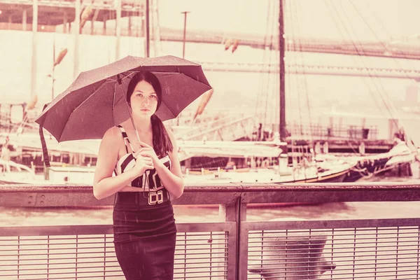 Raining day - grainy, foggy, drizzly feel. American Woman wearing patterned crop bra top, black fitted skirt, holding umbrella, traveling in New York. Boats on background.