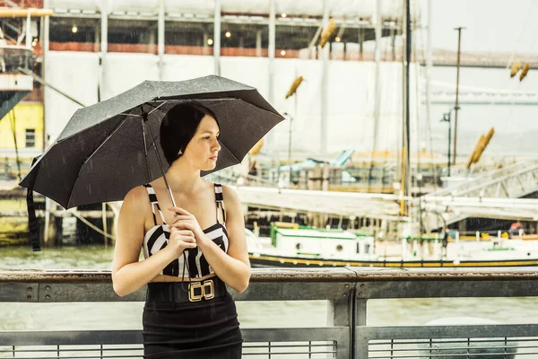 Raining day - grainy, foggy, drizzly feel. American Woman wearing patterned crop bra top, black fitted skirt, holding umbrella, traveling in New York. Boats on background.