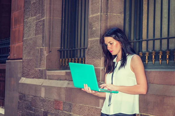 American female college student with long black hair studying in New York in summer, wearing white sleeveless shirt, standing against vintage wall on street, reading, working on laptop computer