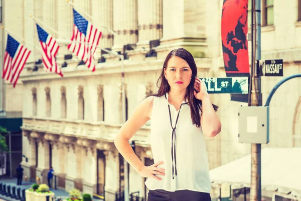American Woman with long black hair traveling in New York in hot summer, wearing white collarless sleeveless shirt, standing on street by vintage building with American flags, relaxing, thinking.