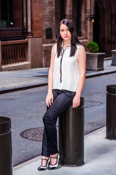 夏にニューヨークを旅する長い黒い髪をしたアメリカの女性は ヴィンテージストリートの金属製の柱の上に座って 白い無色の袖のないシャツ 黒いパンツ ヒールを着て — ストック写真