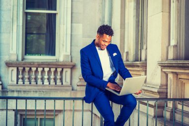 New York 'ta okuyan Afro-Amerikan üniversite öğrencisi, mavi takım elbiseli, beyaz atlet giyen, kampüsteki klasik ofis binasında parmaklıklar üzerinde oturan, aşağıya bakan, dizüstü bilgisayarla çalışan, gülümseyen