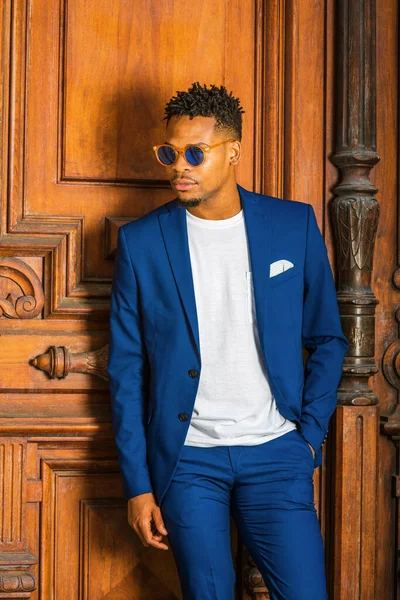 African American Businessman working in New York. Wearing blue suit, unbuttoned, white T shirt, blue sunglasses, college student with little goatee, standing by vintage doorway, looking down, thinking.