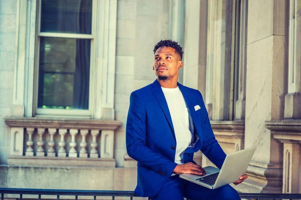 African American college student studying in New York, wearing blue suit, white T shirt, sitting on railing in vintage office building on campus, holding laptop computer, looking up, thinking