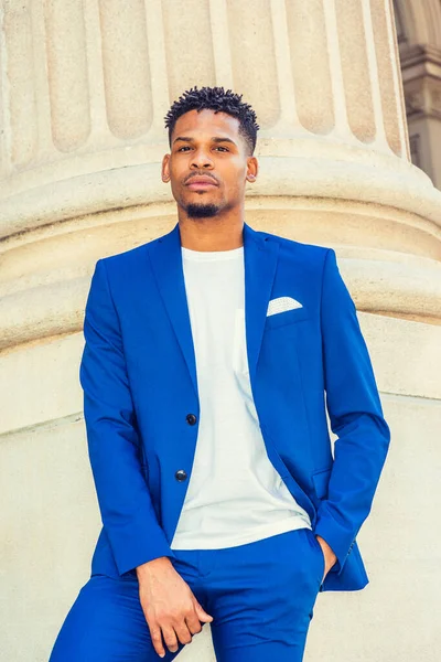 African American Businessman working in New York. Wearing blue suit, unbuttoned, white T shirt, black college student with little goatee, standing by columns on campus, thinking, looking forward