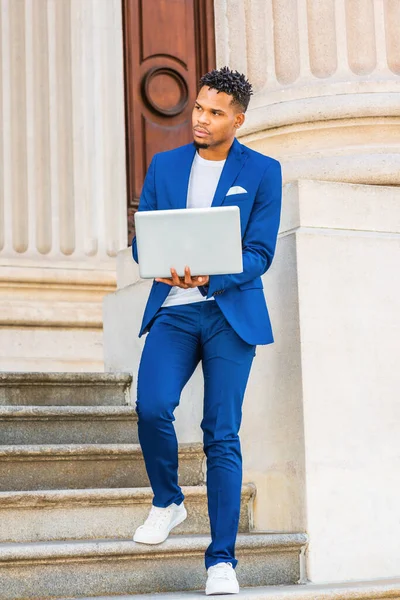 African American college student studying in New York, wearing blue suit, white sneakers, standing on stairs of vintage office building on campus, working on laptop computer, looking away, thinking