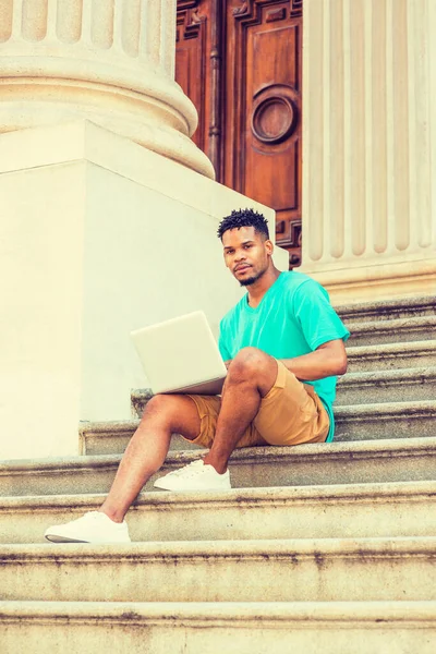 African American college student studying in New York, wearing green V neck T shirt, yellow brown shorts, white sneakers, sitting on stairs of office building on campus, working on laptop computer