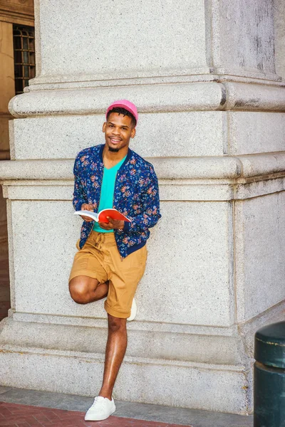 African American college student studying in New York, wearing blue patterned jacket, yellow brown shorts, white sneakers, pink cap, standing against column on campus, reading red book, thinking