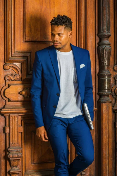 African American Businessman working in New York. Wearing blue suit, white T shirt, college student with little goatee, standing by vintage library door on campus, carrying laptop computer, thinking