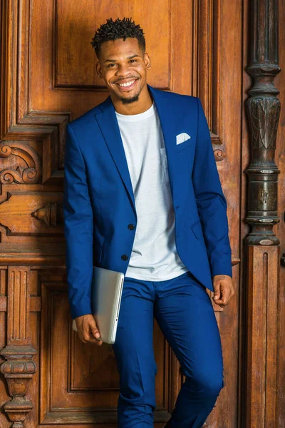 African American Businessman working in New York. Wearing blue suit, white T shirt, college student with little goatee, standing by vintage library door on campus, carrying laptop computer, smiling