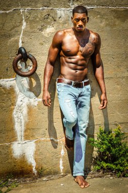Summer Heat. Masculine black man wearing blue jeans, barefoot, half naked, standing against wall with metal ring, under sunshine, looking forward