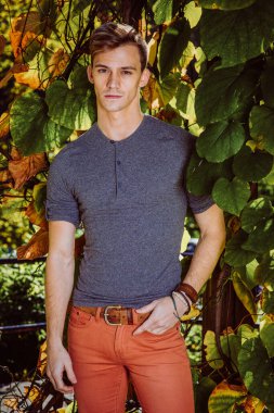 Sonbahar. Gri uzun kollu, yuvarlak taçlı Henley gömlekli, kırmızı kotlu, gün ışığıyla yeşil bitkilerin yanında duran, ileriye bakan genç bir adam.