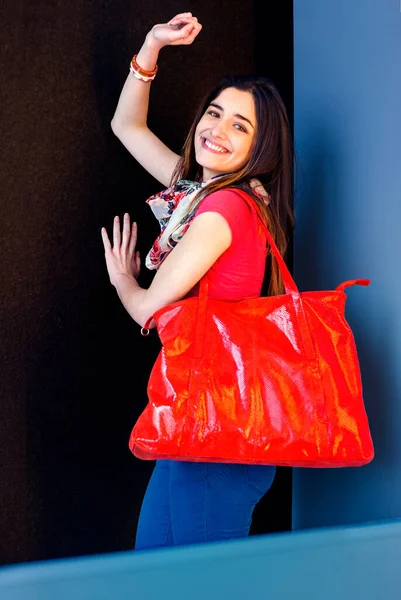 Happy woman with big red bag. Young woman wearing red short sleeve shirt, blue pants, light weight scarf, shoulder carrying big red leather shopping bag, smiling, looking back.