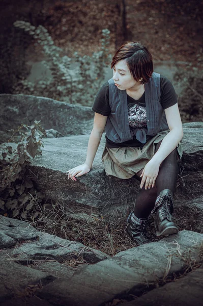 Alone. Young Chinese Girl wearing sleeveless long blouse with black T shirt, black leggings and boots, sitting on rocks, looking around, thinking. Black and White Photograph