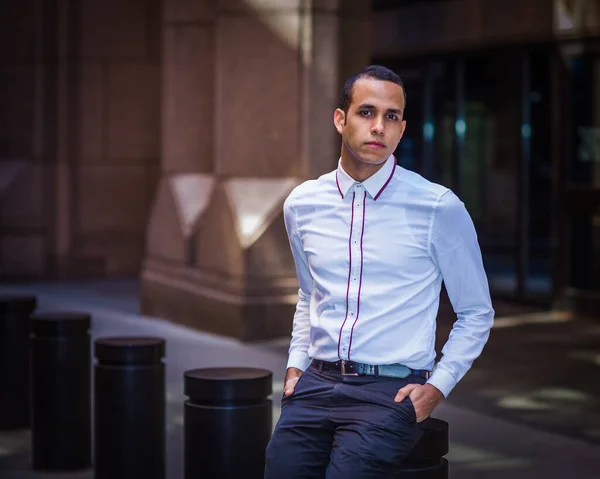 Young Man wearing white shirt, two hands putting in pockets, sitting on metal pillar on old street in New York City, relaxing, thinkin