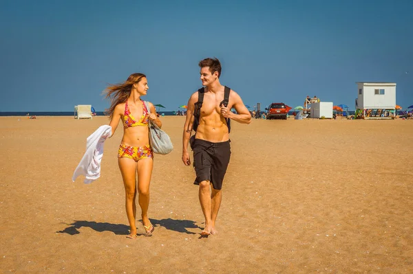 Go Back from Beach. Girl wearing a red, yellow patterned two piece bikini bathing suit, carrying a shoulder bag, a towel, guy wearing a black bathing suit, carrying a back bag, talking, walking.