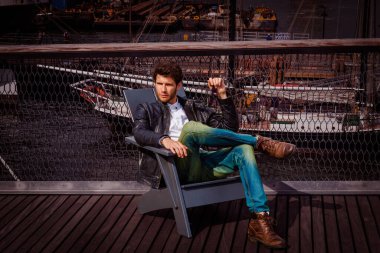 Siyah deri ceket, mavi kot pantolon, kahverengi çizme, güneş gözlüğü takan, New York 'ta güvertede oturan, dinlenen sakallı genç adam. Arka planda tekneler