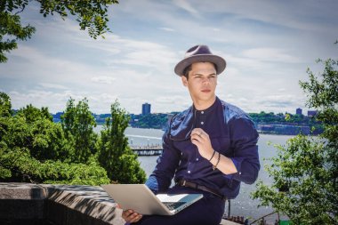 Genç adam, mavi yakalı gömlekli, Fedora şapkalı, güneş gözlüklü, New York 'ta Hudson Nehri' nin kenarında oturuyor, okuyor, dizüstü bilgisayarla çalışıyor.