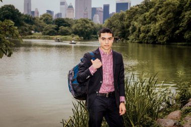 Genç bir üniversite öğrencisi, siyah ceket giyiyor, gömleğinin altında kırmızı, beyaz desenli, omuz çantası taşıyor, New York 'ta gölün kenarında duruyor, ileriye bakıyor.