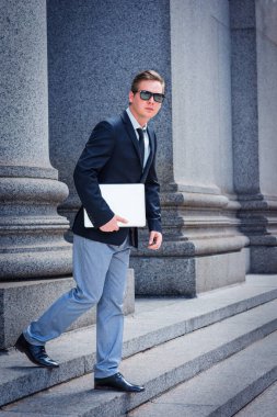Genç adam, siyah ceket, kravat, gri pantolon, deri ayakkabı, güneş gözlüğü, dizüstü bilgisayar taşıyor, New York 'taki ofis binasından merdivenlerden iniyor.