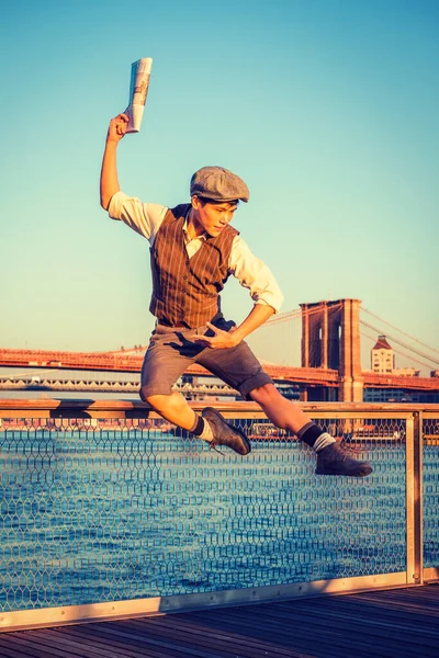 Happy Newsboy in New York City. Young Asian American boy wearing newsboy cap, shirt, vest, pants, leather boot shoes, raising arm, holding newspaper, jumping into air at harbor in sunset, dancing.