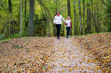 İki kız kardeş el ele tutuşmuş, açık renk ceketler giymişler, sonbaharda ormanda yürüyorlar.