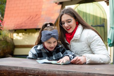 İki kız kardeş bahçedeki ahşap bir masada oturmuş haritaya bakıyor, konuşuyor ve gülüyorlar.